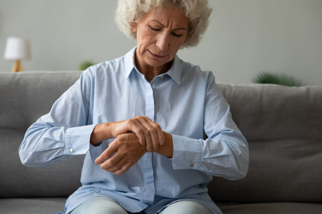 Mulher idosa sentindo fraqueza muscular e dor nas articulações das mãos enquanto está sentada no sofá.