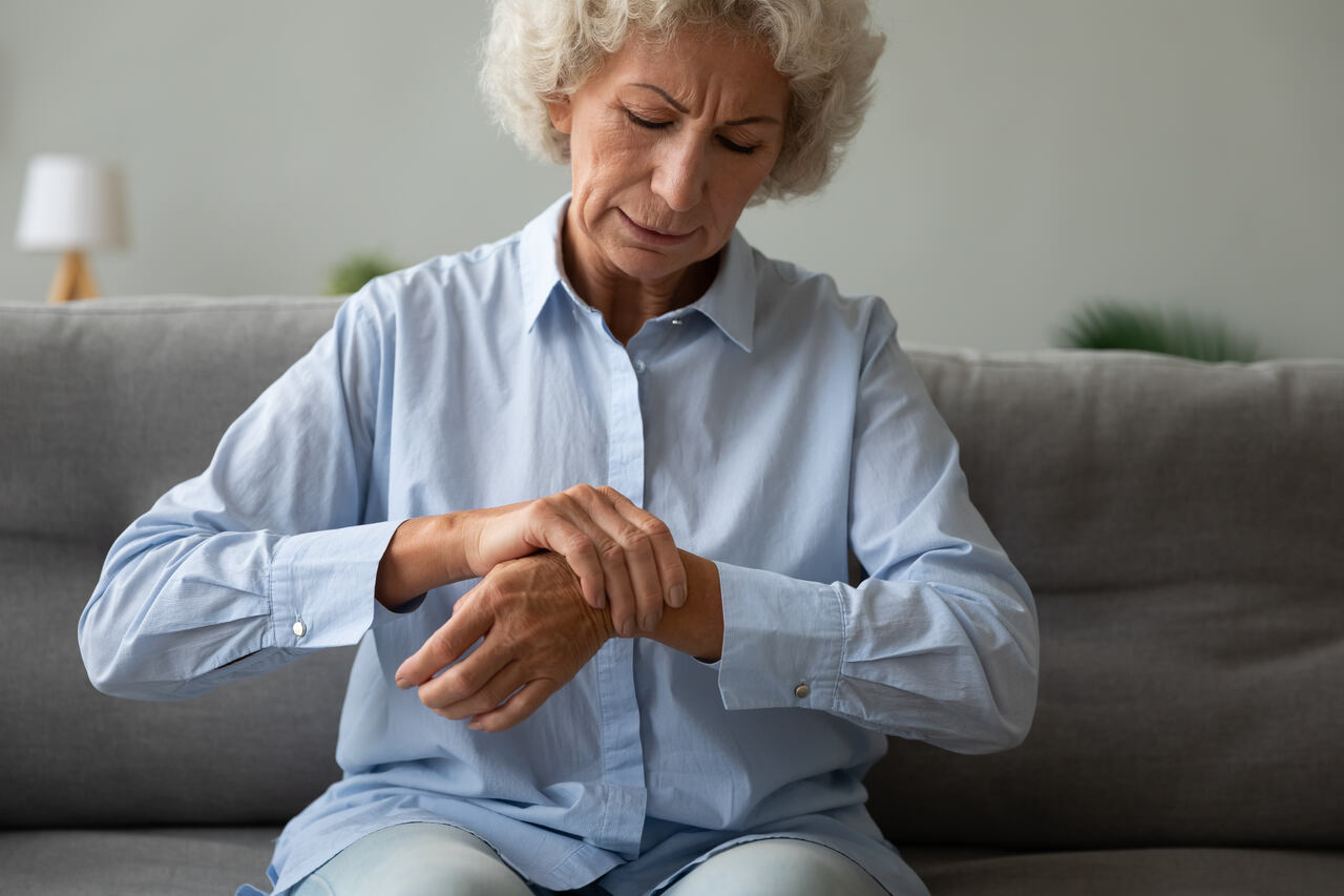 Mulher idosa sentindo fraqueza muscular e dor nas articulações das mãos enquanto está sentada no sofá.