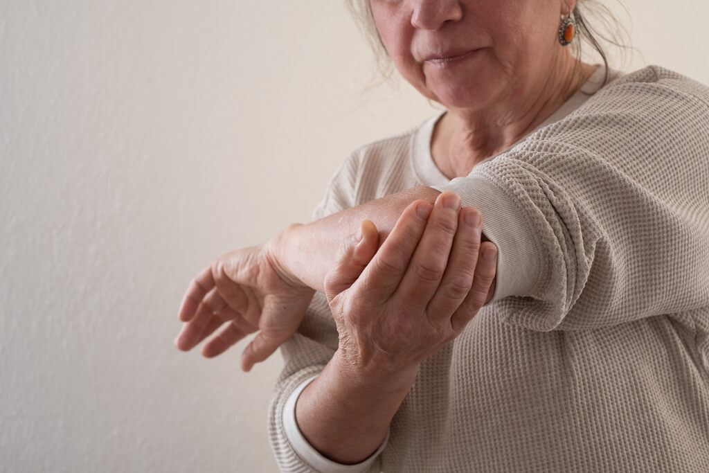 Imagem em close de uma mulher idosa segurando o próprio cotovelo esquerdo com a mão direita, sugerindo dor ou desconforto articular. Ela veste uma blusa de malha bege texturizada. O fundo é neutro e de cor clara, focando a atenção no gesto de cuidado com a articulação.
