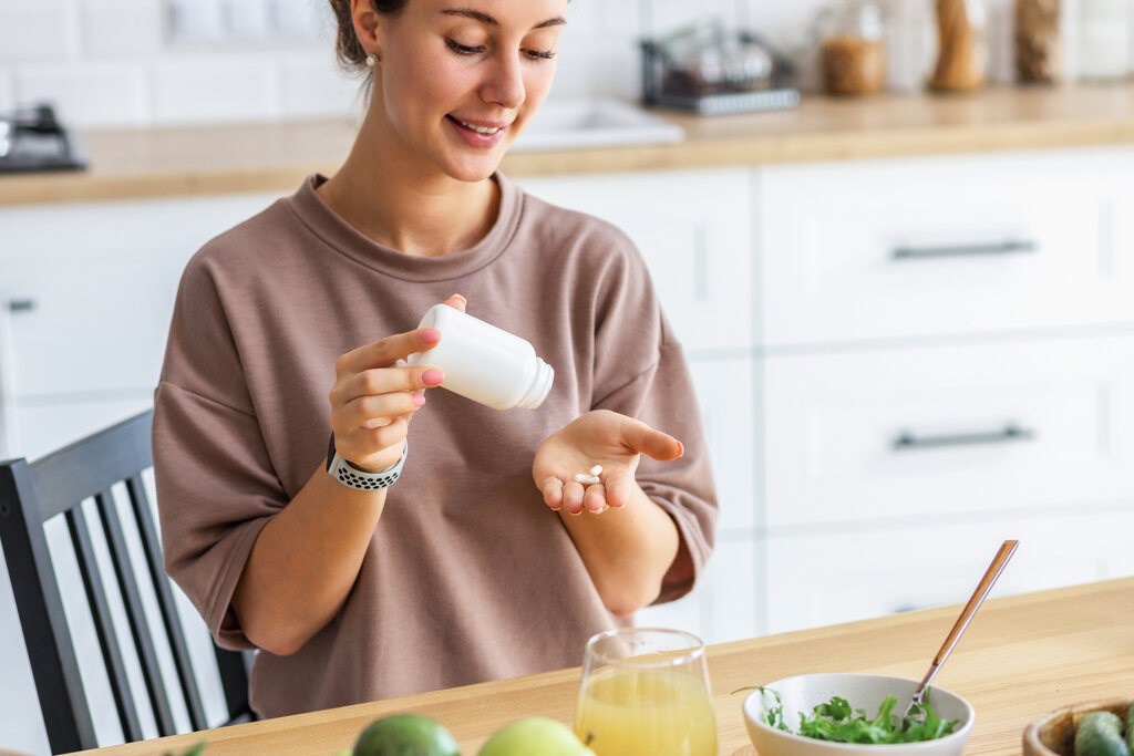 Mulher jovem sorridente na cozinha despejando cápsulas de suplemento de cálcio de um frasco branco na mão.